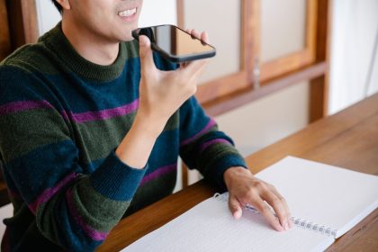 Homem sorrindo usa comandos de voz no celular sentado à mesa em casa.